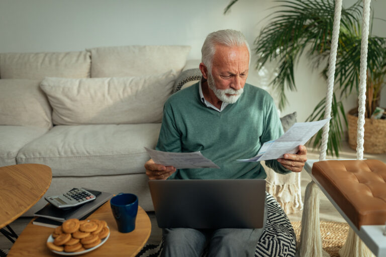 Senior man enjoying in the comfort of the living room, going through paperwork and using laptop