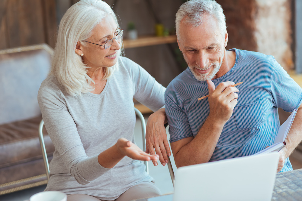 Positive smiling aged couple sitting at the table and browsing the web while having a pleasant talk about financial planning