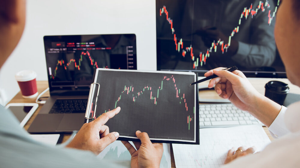 Two people looking at monitors displaying financial stock graph report information.